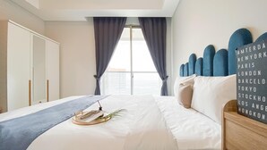 A hotel room with a large bed, a view of the city through the window, a decorative headboard, and a sign listing cities. - Brand New And Homey Studio At Gold Coast Apartment Pik (Jakarta)