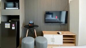 A modern hotel room with a flat-screen TV mounted on the wall, a microwave on the left, a refrigerator with a water bottle holder, a small round table with a bottle, and a wooden cabinet with a laptop and mouse. - Brand New And Homey Studio At Gold Coast Apartment Pik (Jakarta)