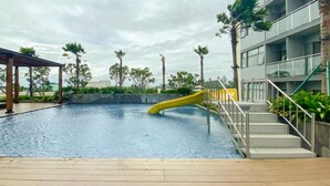 A swimming pool with a yellow slide, surrounded by palm trees and a multi-story building. - Modern And Comfy Studio At Daan Mogot City Apartment (Jakarta)