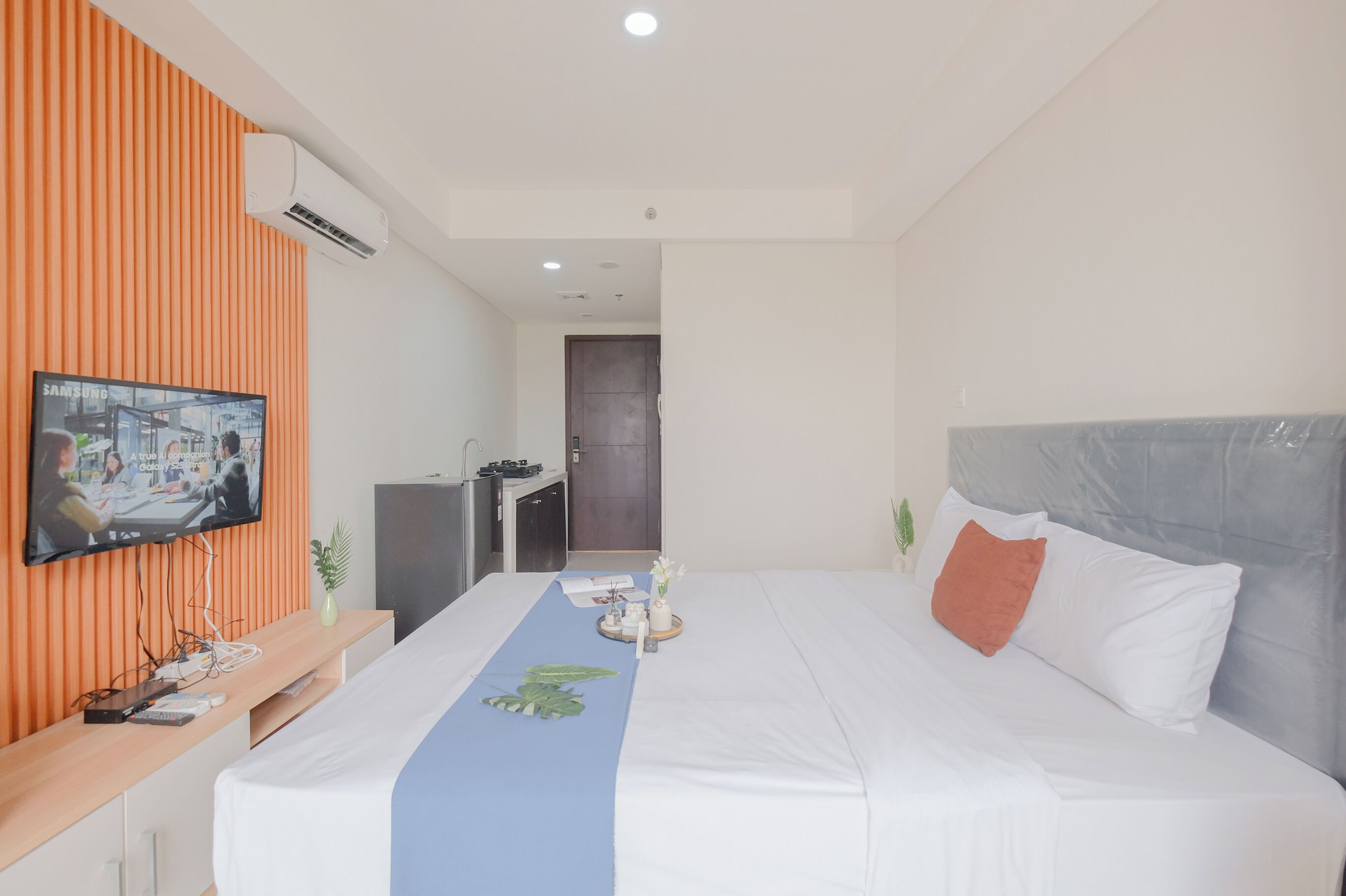 Ein Hotelzimmer mit Bett, einem an einer orangefarbenen Wand montierten Fernseher, einem Schreibtisch mit Computer und einer Tür im Hintergrund.
