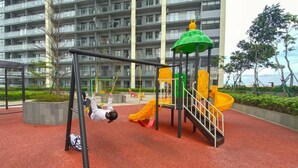 A playground with swings and a slide, surrounded by a red play area and a multi-story building in the background. - Modern And Comfy Studio At Daan Mogot City Apartment (Jakarta)