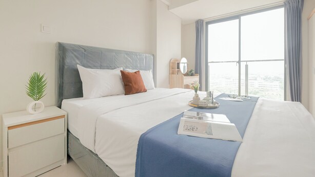 A neatly made bed with a blue bedspread, white pillows, and a brown accent pillow. There is a bedside table with a plant and a small white object. A window with curtains is visible, and a nightstand with a lamp is also present. - Modern And Comfy Studio At Daan Mogot City Apartment (Jakarta)