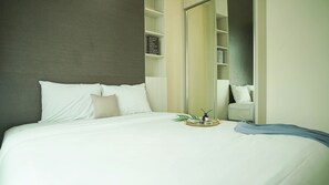 A neatly made bed with white linens and a beige pillow, a bedside table with a plant and a small tray, and a large mirror reflecting the room.