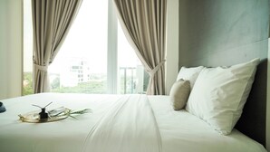A neatly made bed with white linens, a bedside table with a glass tray, and a view of a building through a window with beige curtains.