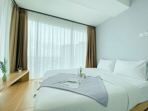 A modern hotel room with a large bed, a bedside table, a window with curtains, and a view of buildings outside. - Comfort And Spacious 2Br Grand Kamala Lagoon Apartment (Bekasi)