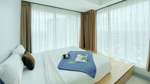 Moderni hotellihuone, jossa on suuri sänky, yöpöytä ja näkymä korkeisiin rakennuksiin ohuiden verhojen läpi.