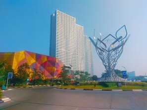 A modern building with a geometric glass facade and a unique metal sculpture in front. - Comfort And Spacious 2Br Grand Kamala Lagoon Apartment (Bekasi)