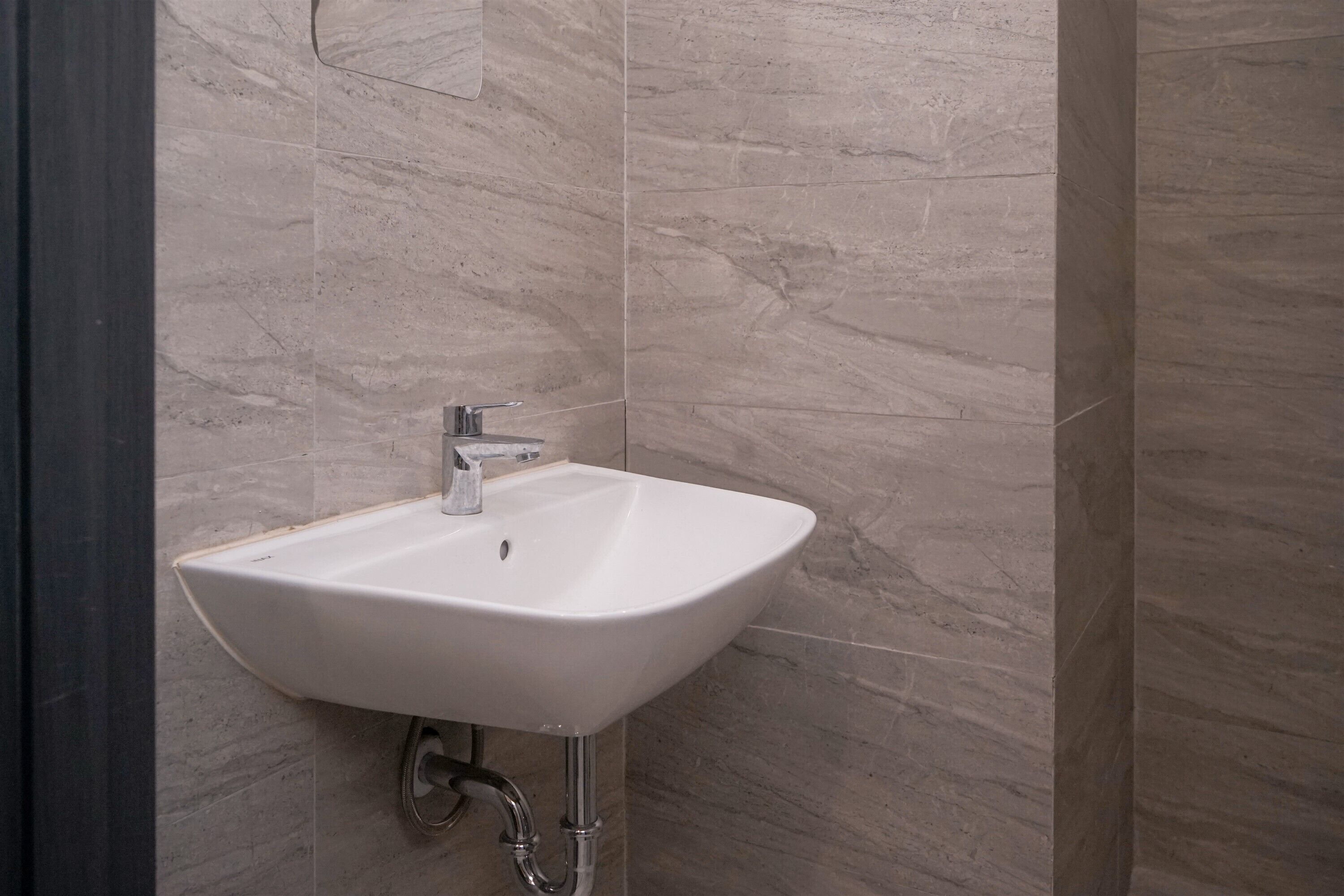 Un baño con lavamanos blanco, paredes de mármol y una tubería a la vista.