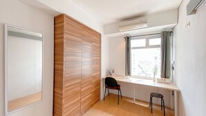 A modern room with a wooden wardrobe, a desk with a chair, a window with curtains, and an air conditioning unit.
