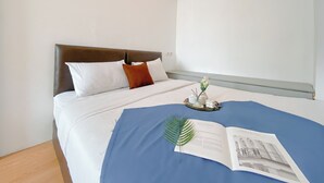 A neatly made bed with white and brown pillows, a blue bedspread, and a small table with a vase and a book.