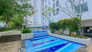 A swimming pool with a tiled deck, surrounded by greenery and a multi-story building in the background. - Cozy Studio Apartment Bassura City (Jakarta)