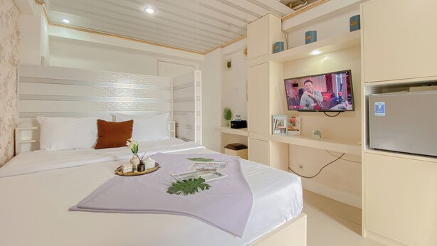 A hotel room with a bed, a television, a refrigerator, and a small desk area. - Cozy Studio Apartment Bassura City (Jakarta)