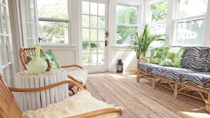A sunlit room with large windows, a wicker sofa, a round table, and a potted plant. - Bellport Inn (Bellport)