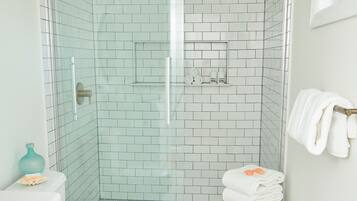 A bathroom with a glass-enclosed shower, white tiled walls, a toilet, a sink with a mirror, and a small wooden stool with towels.