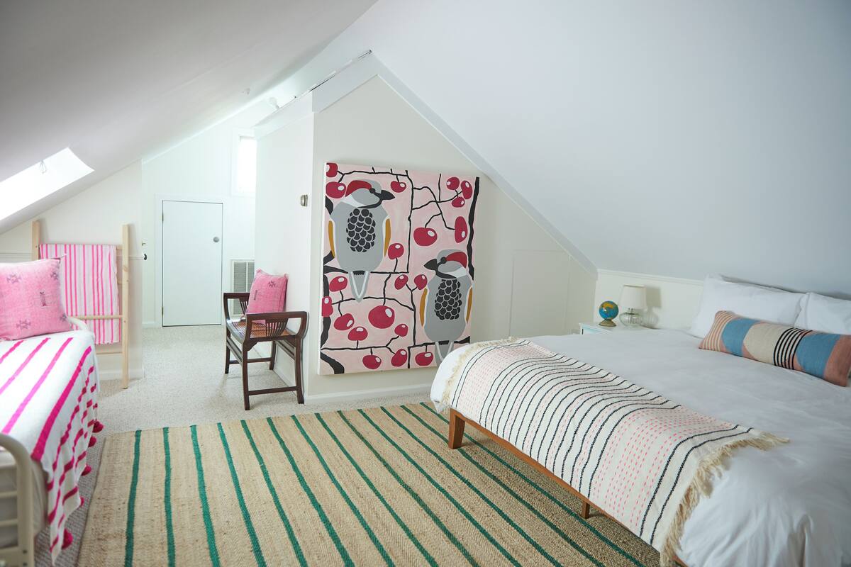 A bedroom with a sloped ceiling, a bed with a striped bedspread, a chair, a desk, and a wall with a fruit pattern.
