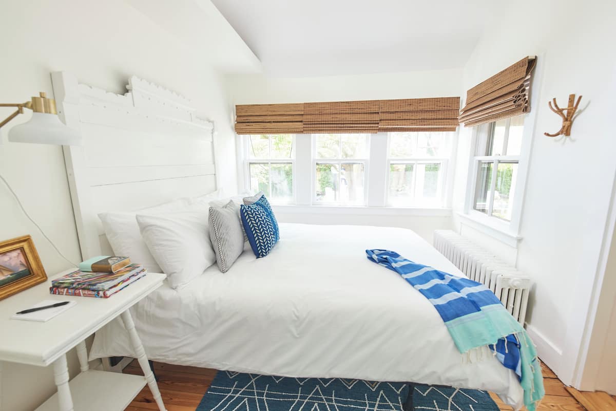 A bedroom with a white bed, a bedside table with books, a lamp, and a view of the outdoors through large windows.