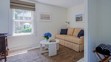 Un salon avec un canapé à rayures, une table basse blanche ornée d’un vase de fleurs, une fenêtre avec des stores et une vue sur une maison à l’extérieur.