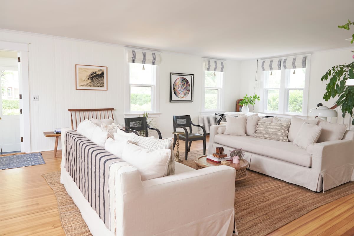 A living room with a sofa, armchairs, a coffee table, and a dining table. There are two windows with curtains, a painting on the wall, and a potted plant.