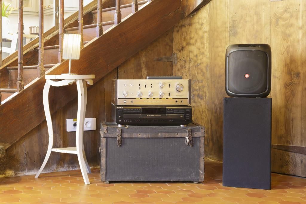 A staircase with a wooden railing, a white chair, a vintage Sony stereo system on a suitcase, and a black speaker on a stand.
