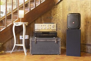 Un escalier avec une rampe en bois, une chaise blanche, une chaîne stéréo Sony vintage posée sur une valise et un haut-parleur noir sur un pied.