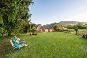 Un espacio al aire libre con césped, una fila de sillones y una mesa, un edificio de piedra con una puerta roja y una montaña al fondo.