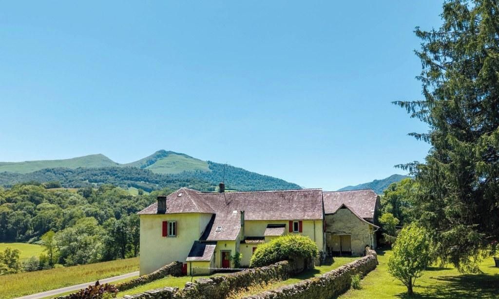 Une maison en pierre à deux étages, dotée d’un toit en pente, entourée d’un mur de pierre et d’un champ herbeux.