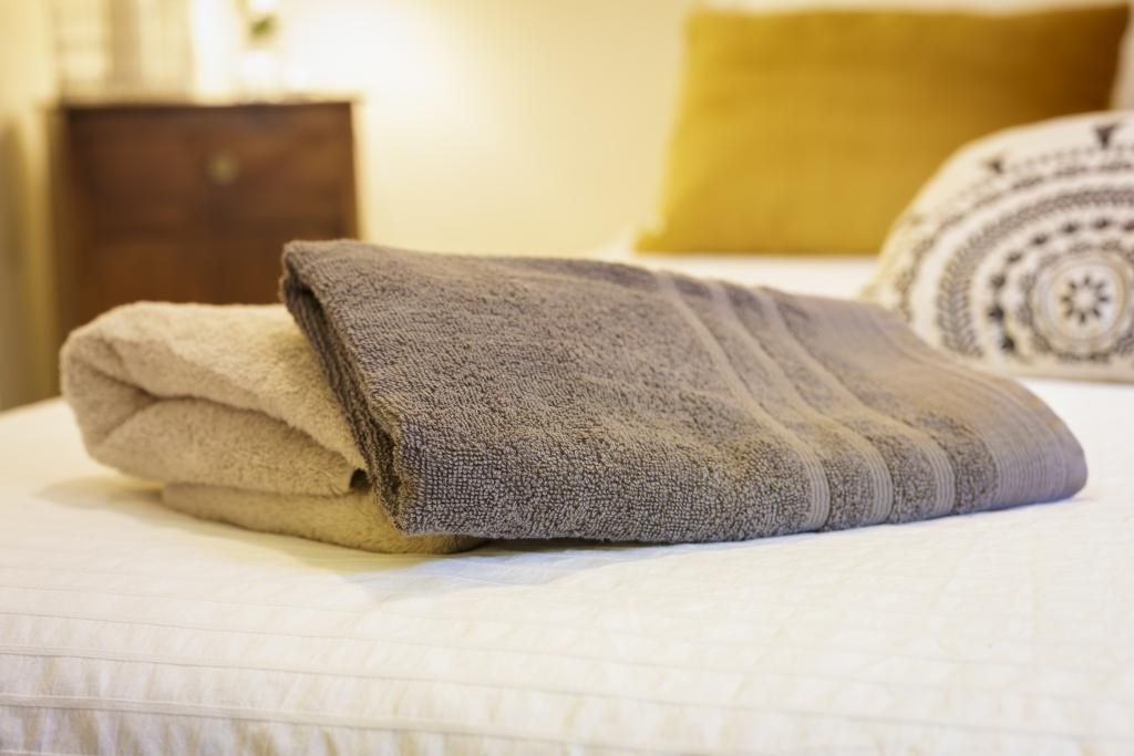 A neatly folded towel on a bed with a patterned pillow and a wooden nightstand in the background.