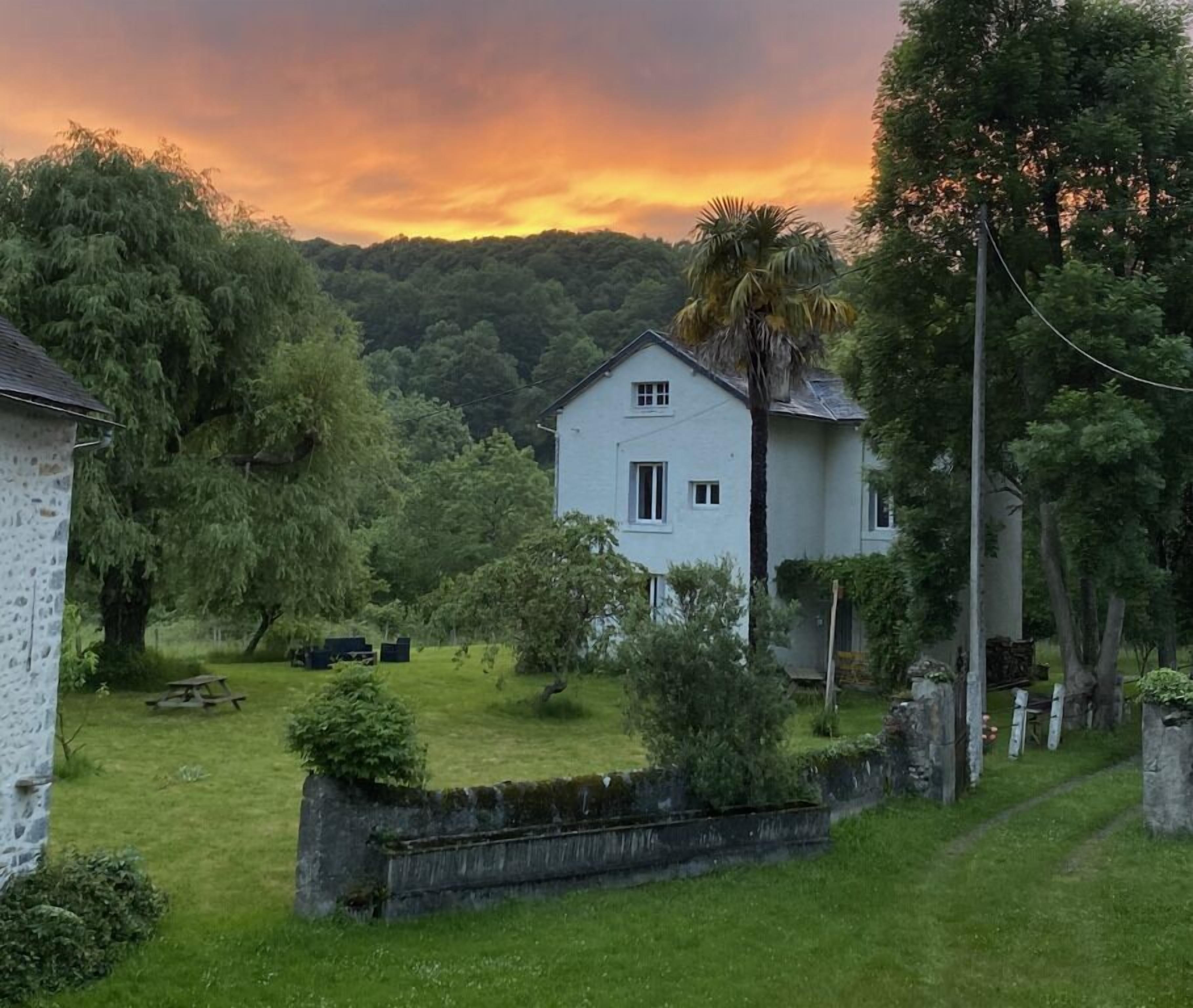 Une maison blanche avec un petit jardin devant, entourée d’arbres et d’un mur de pierres.