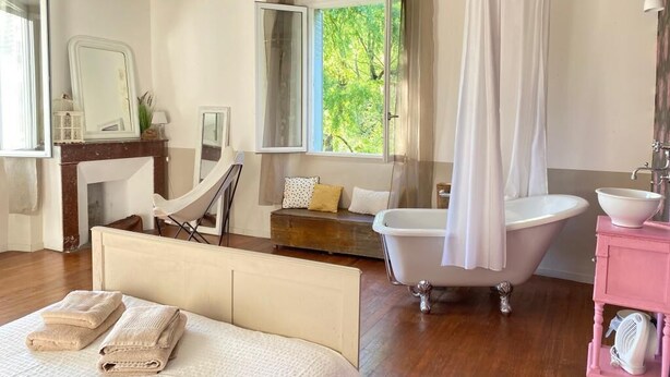 Une chambre avec un grand lit, une baignoire blanche, une coiffeuse avec un meuble rose et une fenêtre donnant sur un paysage verdoyant.