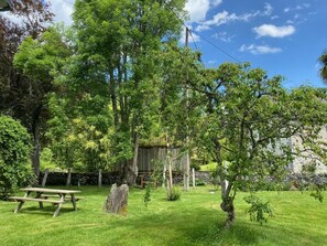 A grassy outdoor area with a picnic table and a stone structure, surrounded by trees and a stone wall.