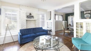 A living room with a blue sofa, a glass coffee table, and a teal armchair. There is a white brick wall with a 'welcome' sign, a floor lamp, and a dining area visible in the background. - Sleeps 12 • Budget-Friendly Downtown Birmingham Crew House (Birmingham)