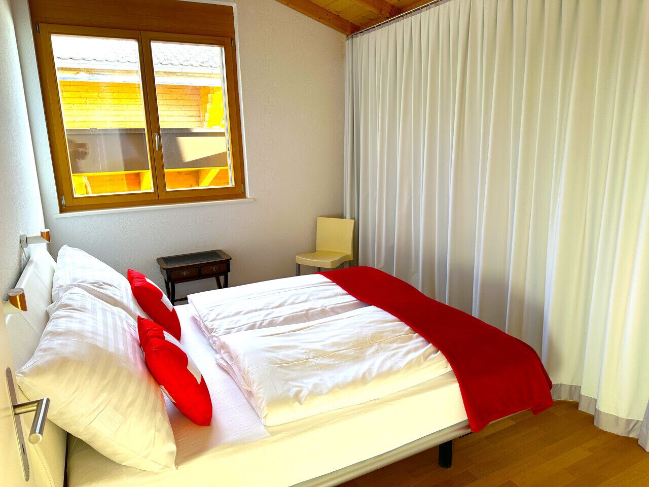 Un lit bien fait, avec du linge de lit blanc et des coussins rouges, un plancher en bois, une fenêtre avec des stores et une chaise près de la fenêtre.