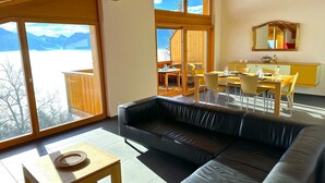 Een moderne woonkamer met een leren bank, een houten eettafel en stoelen. Grote ramen bieden uitzicht op bergen en een houten terras.