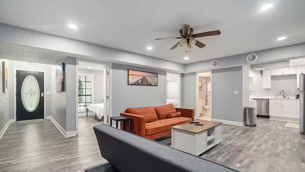 A modern living room with a grey sofa, a coffee table, and a ceiling fan. There is a kitchen area with white cabinets and a dining table. - Cozy Apartment in Lilburn - Unit A + Unit C (Lilburn)