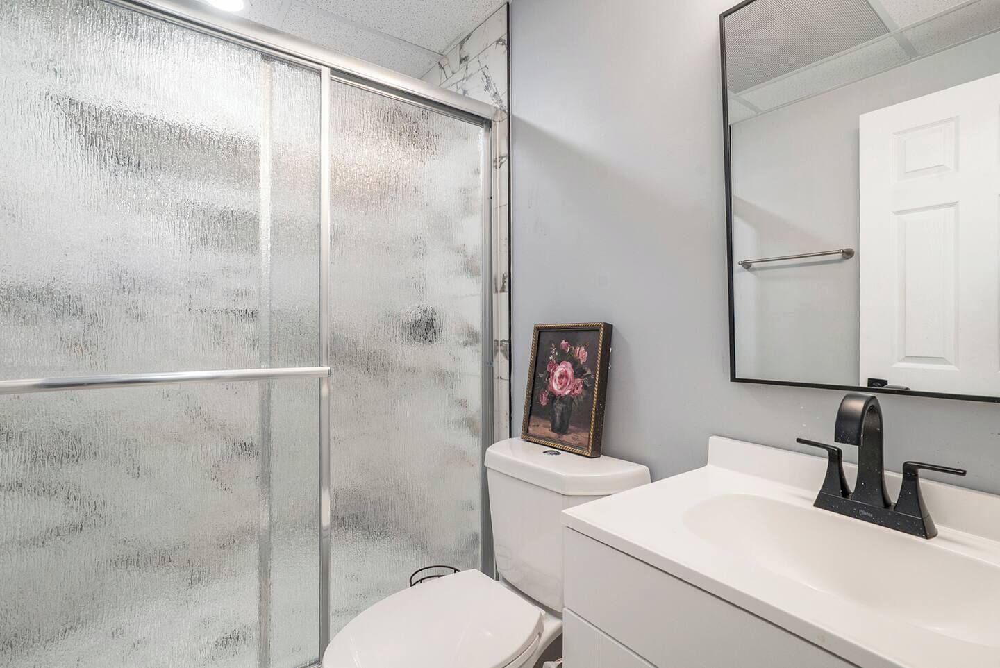 A bathroom with a glass shower enclosure, a toilet, a sink with a black faucet, and a framed picture on the wall.
