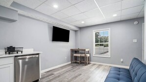 A modern kitchen with a stainless steel refrigerator, a built-in oven, and a flat-screen TV mounted on the wall. There is a wooden dining table with two chairs and a blue sofa in the living area. - Cozy Apartment in Lilburn - Unit A + Unit C (Lilburn)