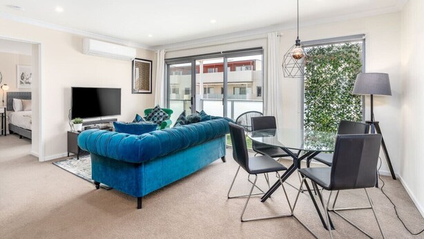 A modern living room with a blue sofa, a flat-screen TV, and a dining area with a glass table and chairs. - Cosy 2BR Close to Light Rail (Harrison)