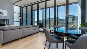 Una sala de estar moderna con un sofá gris, una mesa de comedor de vidrio y sillas. Amplias ventanas ofrecen vistas panorámicas de la ciudad.