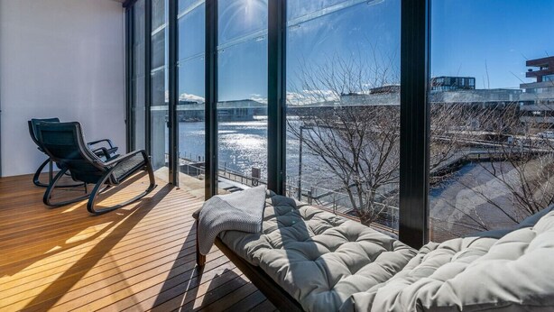 Una habitación moderna con un ventanal grande, una terraza de madera, un sofá y un sillón mecedor.