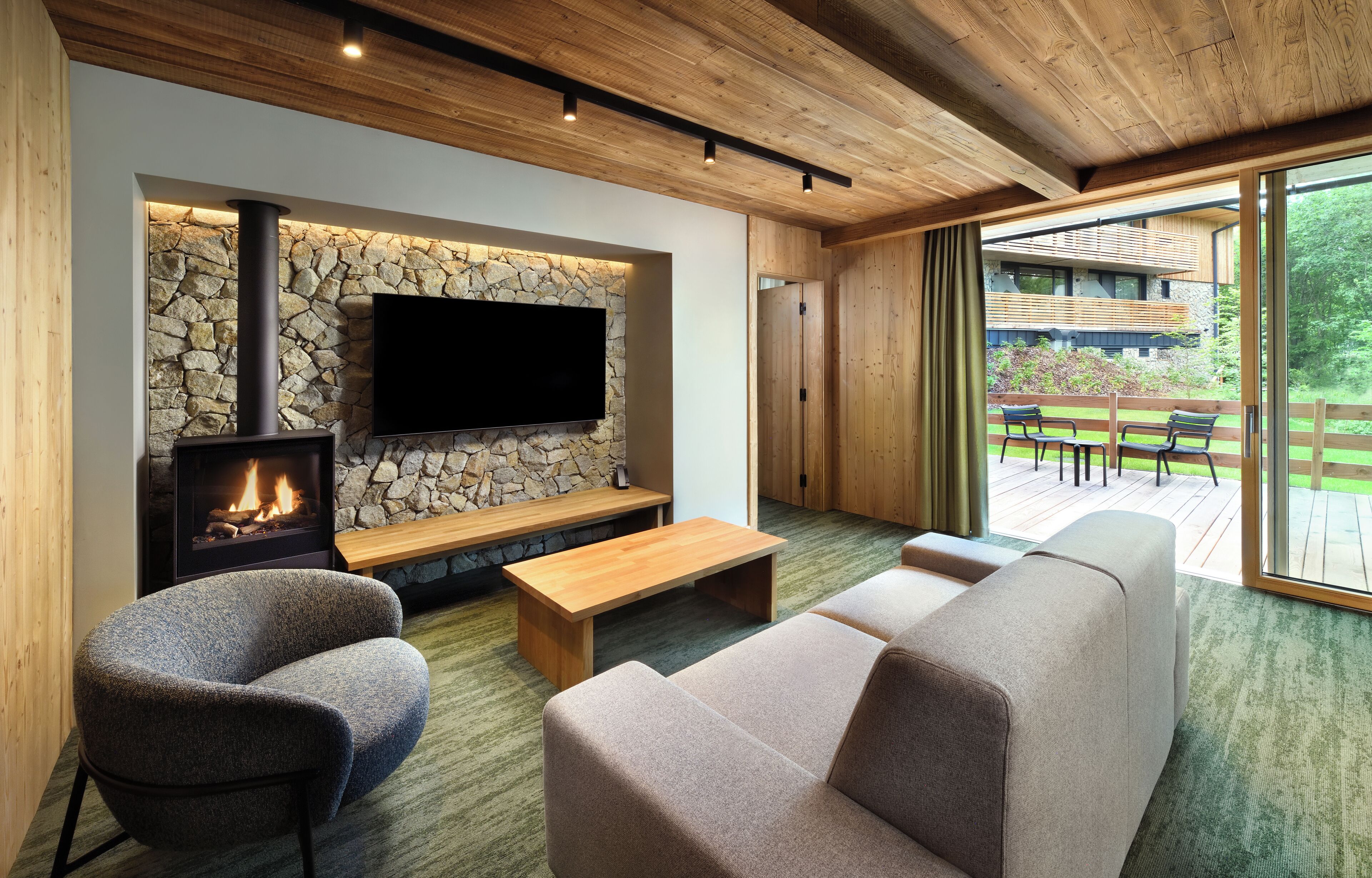 A modern living room with a stone wall, a fireplace, a flat-screen TV, and a large window overlooking a deck.