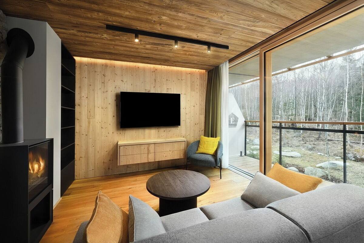 Una sala de estar moderna con chimenea, un televisor grande montado en la pared y vistas a un bosque a través de amplios ventanales.