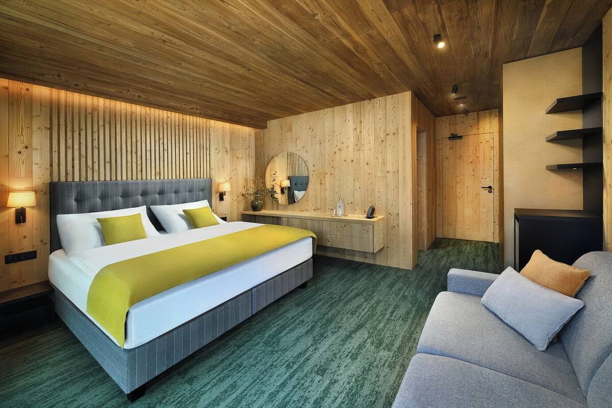 Una habitación de hotel moderna con una cama grande, un sofá gris, paneles de madera, un espejo redondo y una mesita de noche pequeña.