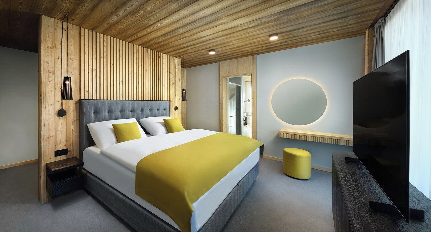 A modern hotel room with a large bed, wooden panel walls, a round mirror, and a flat-screen TV.
