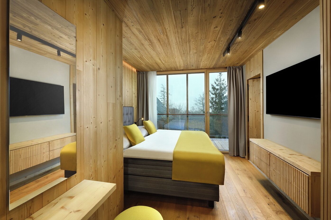 Un dormitorio moderno con una cama grande, paneles de madera, un televisor y vistas a árboles a través de amplios ventanales.