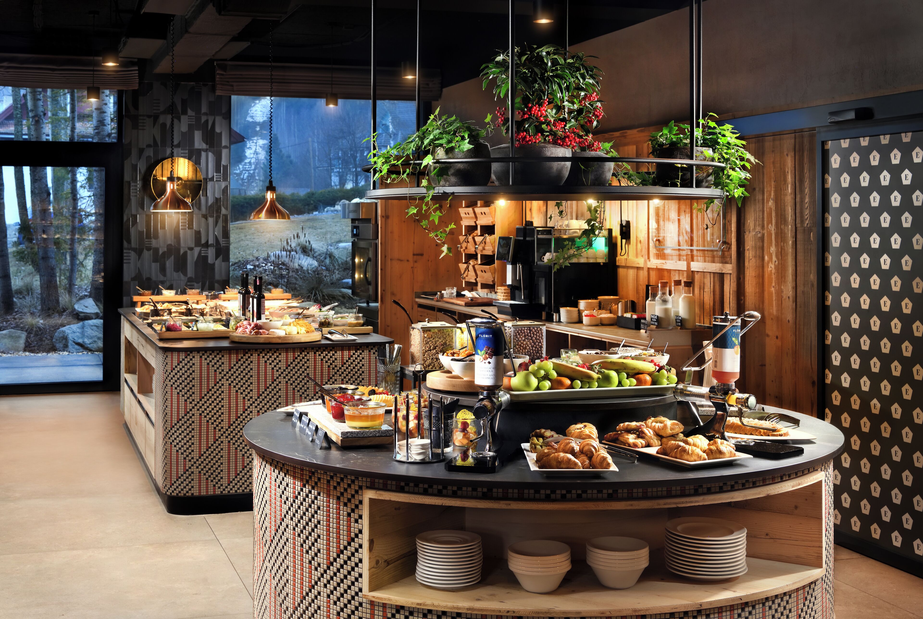 A buffet area with a variety of food items, including fruits, pastries, and beverages, arranged on a counter with hanging plants.