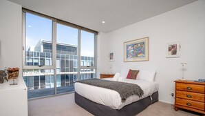 A modern bedroom with a large bed, a dresser, and a view of buildings through a sliding glass door.