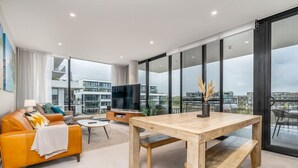 Un moderno salón con una mesa de comedor de madera, un sofá, un televisor y grandes ventanales que ofrecen vistas a la ciudad.