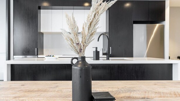 Una cocina moderna con gabinetes oscuros, una encimera blanca y un florero con plantas secas sobre una mesa de madera.