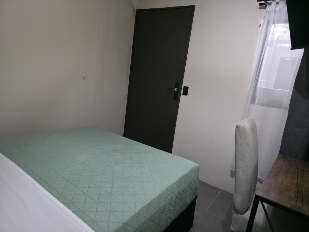 A small, single-bed room with a plain door, a chair, and a window with curtains.