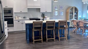 Una cucina moderna con mobili bianchi, una grande isola con sgabelli da bar e un'area pranzo con sedie in vimini.